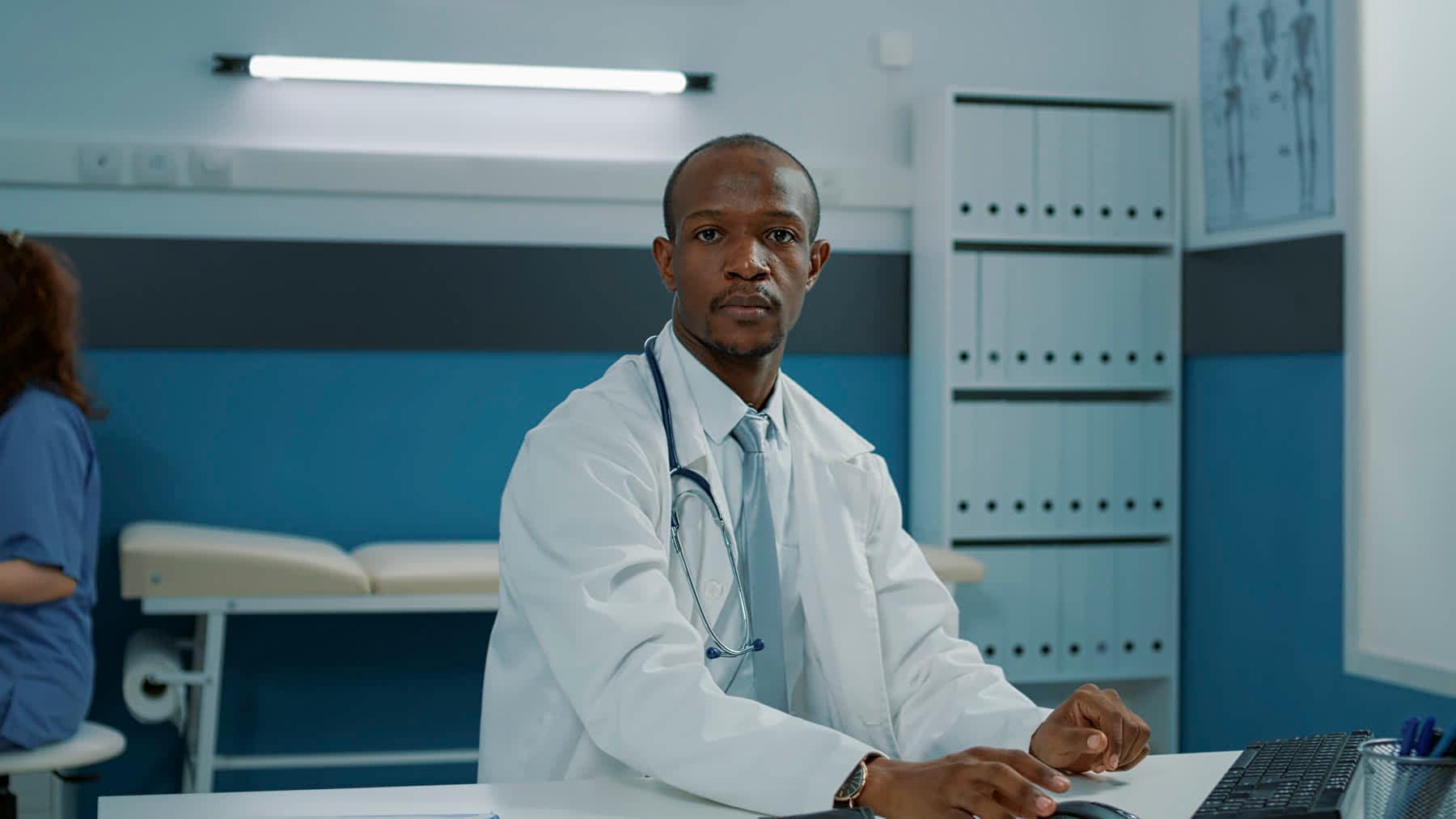 A Black doctor in a white coat and tie, working at a desktop computer in his consulting room