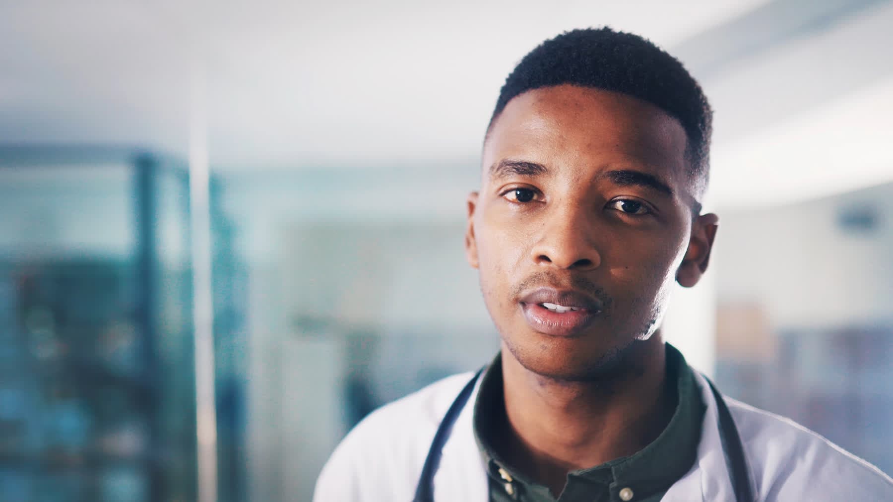 A young Black doctor in a white coat and stethoscope, looking steadily at the camera in a bright hospital lobby