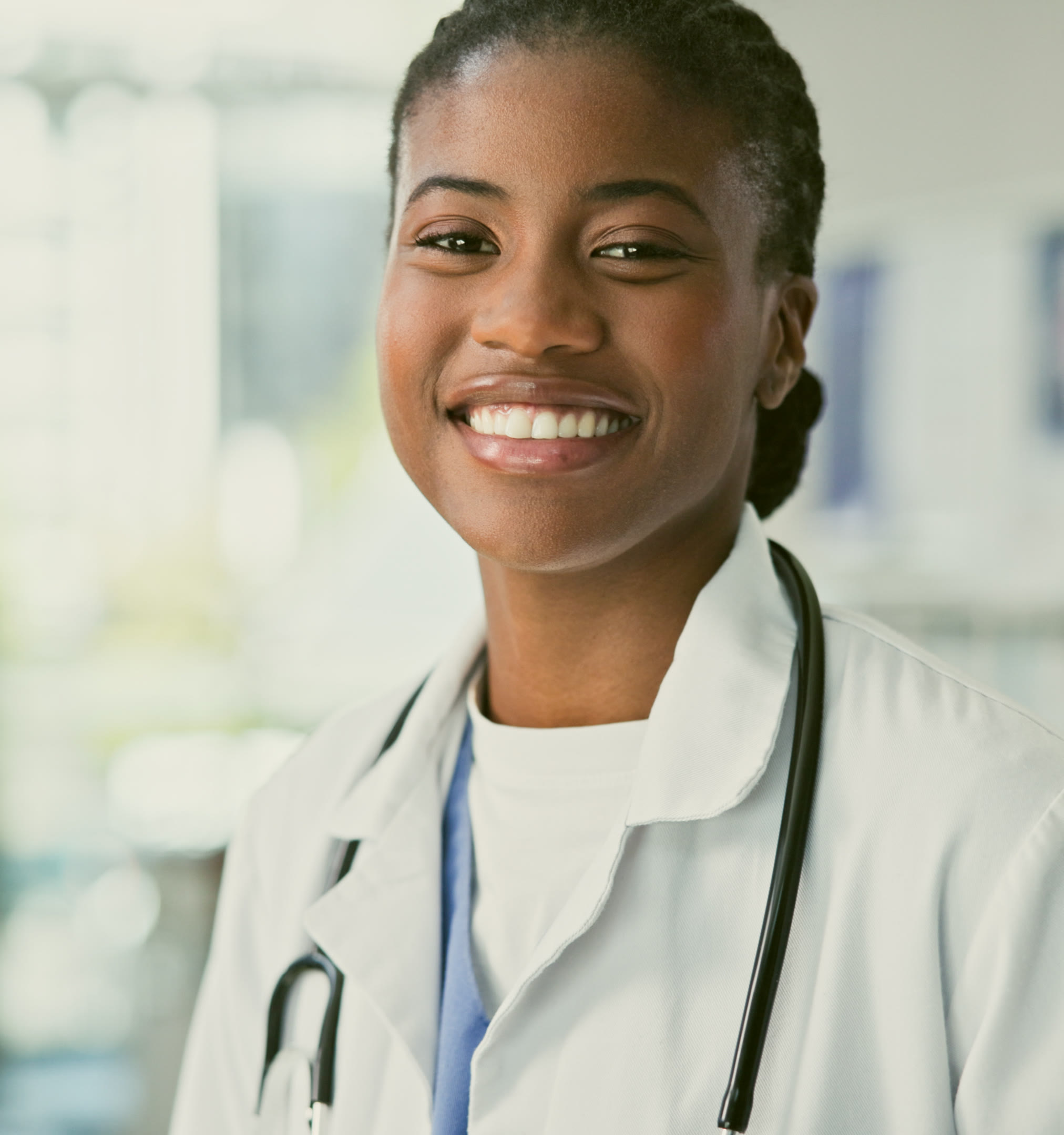 A smiling Black woman GP in a white coat and stethoscope, standing in a bright hospital corridor